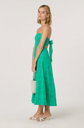 A woman is wearing a strapless, turquoise dress with eyelet embroidery. The dress has a fitted bodice and a high waist, with a matching sash tied in a bow on her left side. The skirt is mid-calf length, flowy, and features a circular eyelet pattern. She is also wearing white high-heeled sandals with thin straps.